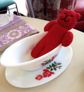 a photograph of a collection of objects including a red knitted teddy bear inside a white china gravy boat with red and green flower decoration