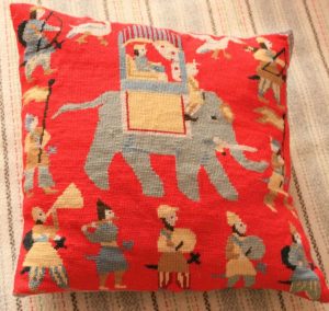photograph of an orange coloured tapestry cushion decorated with people and an elephant sewn in shades of grey, cream and brown