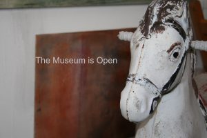 the head of a white vintage rocking horse in front of a blurred orange painting with 'The Museum is Open' written in small white letters