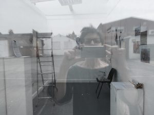 multiple layered image of a woman with short hair and glasses taking a photograph with her camera through a window. The interior of a white room with paintings and a ladder can be seen